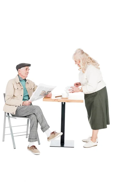 Retired woman looking at husband with newspaper while pouring tea in cup isolated on white — Stock Photo