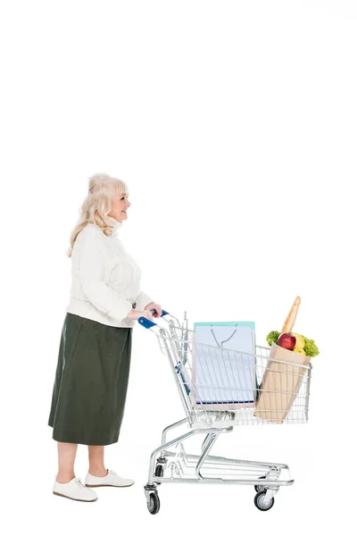Cheerful retired woman walking with shopping cart with shopping bags and paper bag with groceries isolated on white — Stock Photo