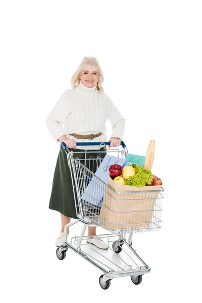Mujer mayor feliz sosteniendo carro de la compra con bolsas de compras y bolsa de papel con comestibles aislados en blanco - foto de stock