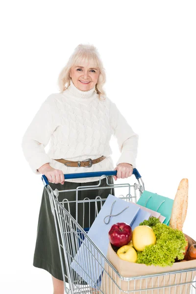 Sonriente mujer mayor sosteniendo carrito de compras con bolsas de compras y bolsa de papel con comestibles aislados en blanco - foto de stock