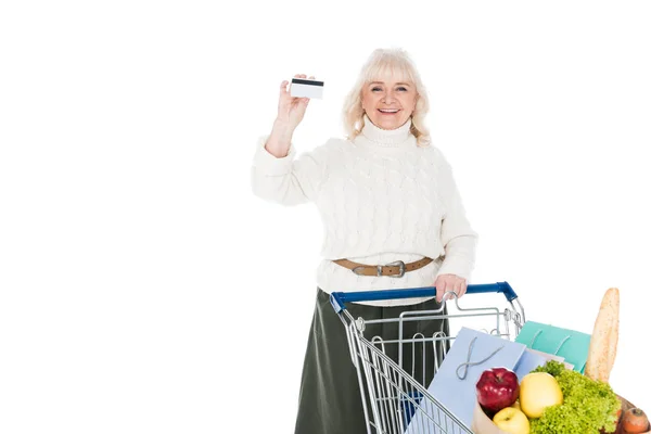 Smiling senior woman holding credit card near shopping trolley isolated on white — Stock Photo