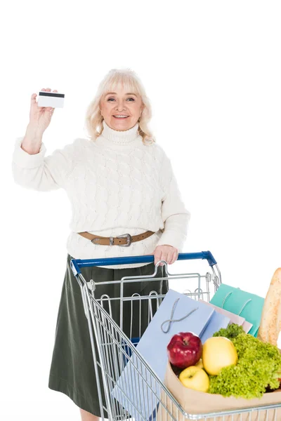 Femme âgée gaie tenant la carte de crédit près du chariot d'achat isolé sur blanc — Photo de stock