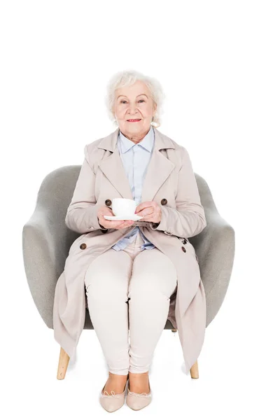 Fröhliche Seniorin sitzt im Sessel mit einer Tasse Tee isoliert auf weiß — Stockfoto