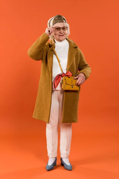 Stylish senior woman touching sunglasses and bag on orange background — Stock Photo