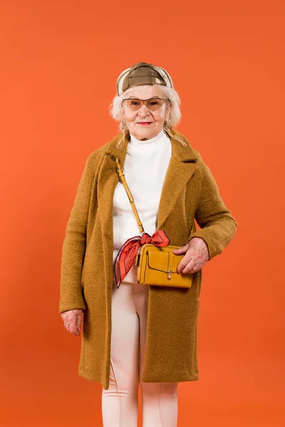 Elegante mujer mayor tocando bolsa aislada en naranja - foto de stock