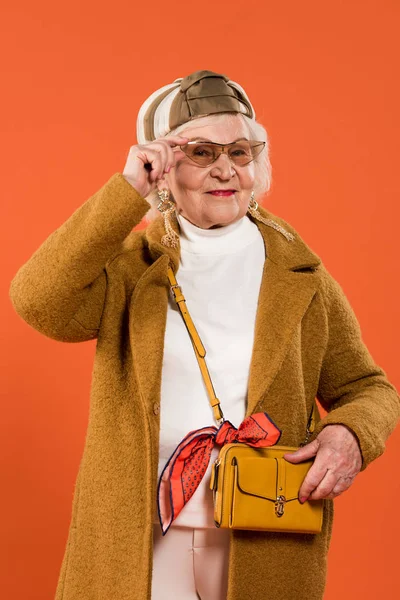 Élégant senior femme touchant lunettes de soleil isolées sur orange — Photo de stock