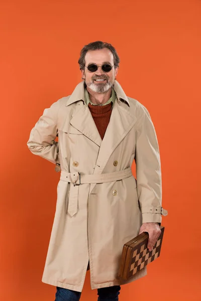 Homem idoso alegre em óculos de sol de pé com tabuleiro de xadrez isolado em laranja — Fotografia de Stock