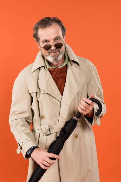 Homme âgé joyeux dans les lunettes de soleil tenant parapluie isolé sur orange — Photo de stock