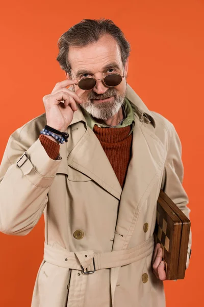 Модный пожилой человек трогает солнцезащитные очки, держа шахматную доску изолированной на оранжевом — стоковое фото