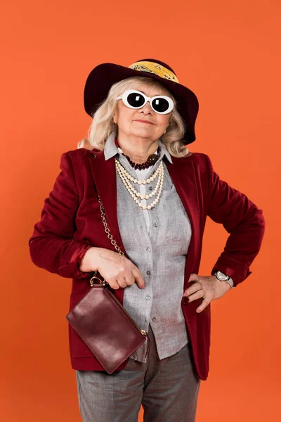 Trendy senior woman in sunglasses and hat smiling isolated on orange — Stock Photo
