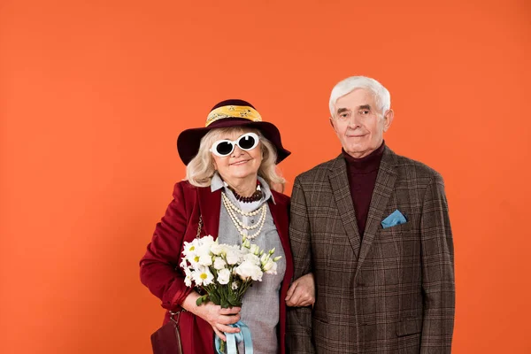 Стильна старша жінка в сонцезахисних окулярах і капелюсі стоїть з чоловіком, тримаючи квіти ізольовані на помаранчевому — стокове фото