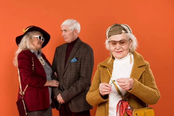 Foyer sélectif de la femme âgée élégante dans les lunettes de soleil tenant fleur avec couple à la retraite sur fond isolé sur orange — Photo de stock