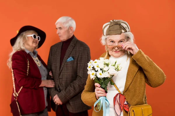 Selective focus of stylish senior woman in sunglasses holding bouquet with retired couple on background isolated on orange — Stock Photo