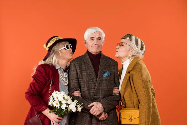 Mujeres mayores de moda de pie entre el hombre jubilado y el envío de besos aislados en naranja - foto de stock