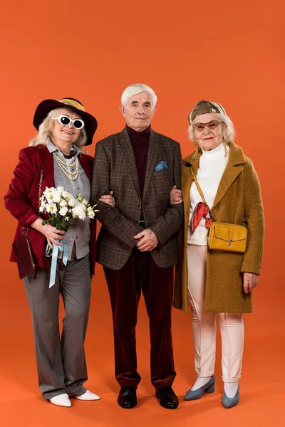 Mujeres mayores con estilo de pie entre el hombre jubilado y la celebración de flores sobre fondo naranja - foto de stock