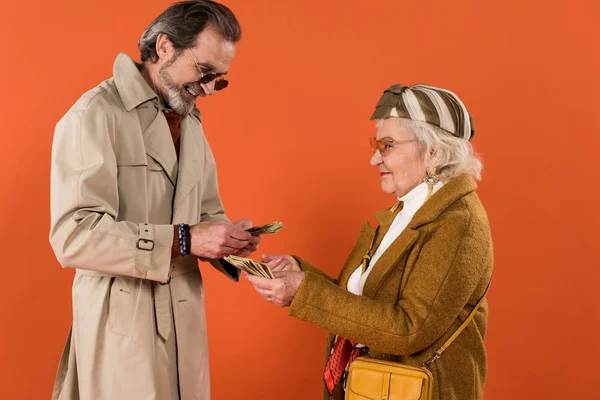 Élégant couple de personnes âgées tenant des billets en dollars dans des mains isolées sur orange — Photo de stock