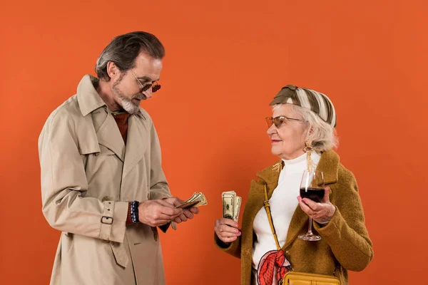 Mulher aposentada feliz segurando notas de dólar e copo de vinho enquanto olha para o homem isolado em laranja — Fotografia de Stock
