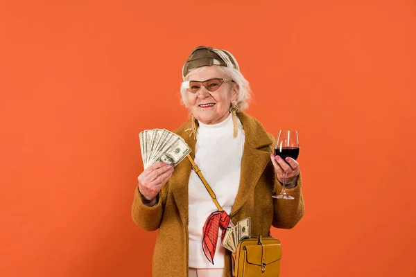 Alegre anciana sosteniendo dinero y copa de vino en manos aisladas en naranja - foto de stock