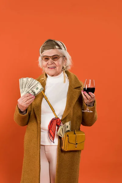 Heureuse femme âgée tenant de l'argent et un verre de vin dans les mains isolées sur orange — Photo de stock