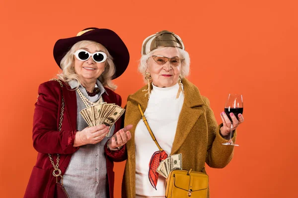 Alegre anciana sosteniendo dinero cerca de amigo con copa de vino en mano aislado en naranja - foto de stock