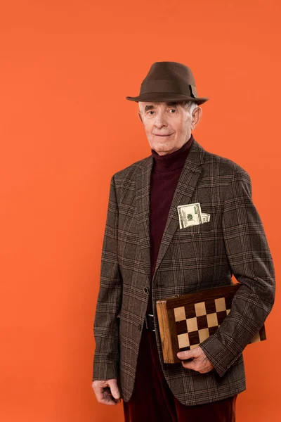Homem sênior na moda em chapéu segurando tabuleiro de xadrez com dinheiro no bolso isolado em laranja — Fotografia de Stock