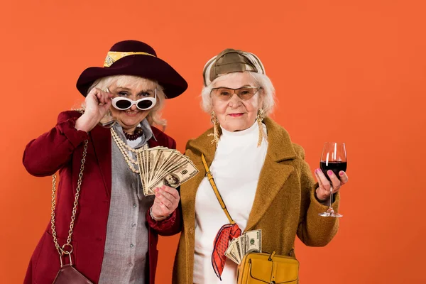 Mujer mayor tocando gafas de sol mientras sostiene el dinero cerca de amigo con copa de vino en la mano aislado en naranja - foto de stock