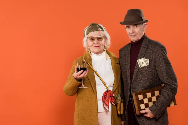 Glückliche Rentnerin hält ein Glas Wein in der Hand, während sie neben einem Mann steht, der Geld in der Tasche hat, isoliert auf Orange — Stockfoto