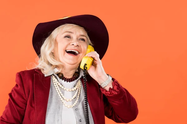 Веселая женщина в шляпе говорит по ретро-телефону изолированы на оранжевый — стоковое фото