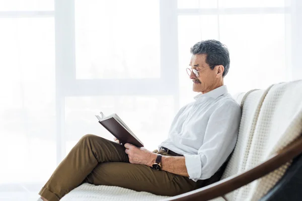 Homme âgé réfléchi livre de lecture tout en étant assis sur le canapé — Photo de stock