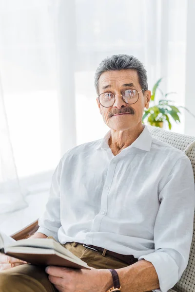 Rentner mit Brille hält Buch in der Hand und lächelt zu Hause — Stockfoto