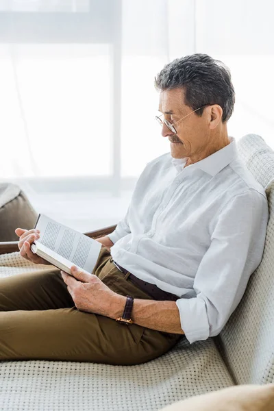 Старший чоловік читає книгу, сидячи на дивані вдома — стокове фото