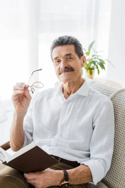 Homme âgé gai tenant livre et lunettes à la maison — Photo de stock