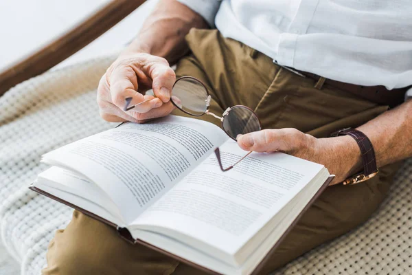 Cropped view of senior man reading book and holding glasses at home — Stock Photo