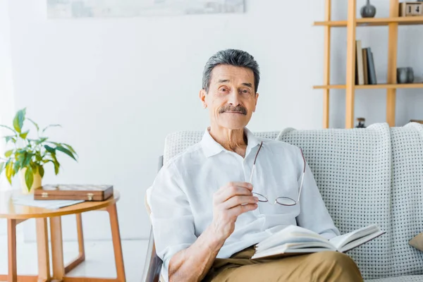 Heureux homme âgé tenant livre et lunettes à la maison — Photo de stock