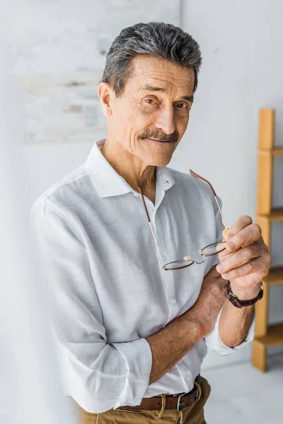 Happy senior man holding glasses and smiling at home — Stock Photo