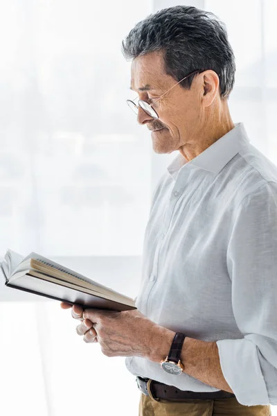 Пенсионер в очках стоя и читая книгу дома — стоковое фото