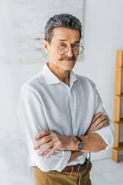 Senior dans des lunettes debout avec les bras croisés à la maison — Photo de stock