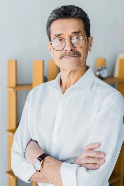 Retired man in glasses standing with crossed arms at home — Stock Photo