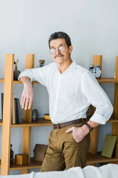 Uomo anziano in occhiali in piedi vicino alla libreria con mano in tasca a casa — Foto stock