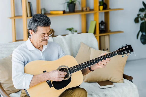 Happy pensioner in glasses playing acoustic guitar at home — Stock Photo