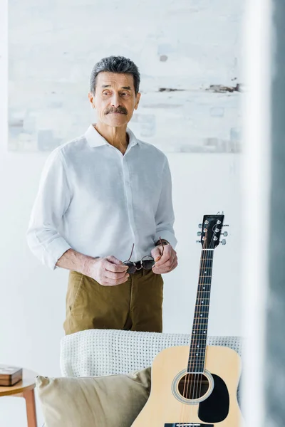 Triste hombre mayor de pie cerca de la guitarra acústica en casa - foto de stock