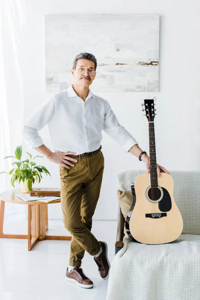 Пенсионер, стоящий со скрещенными ногами возле акустической гитары в гостиной — стоковое фото