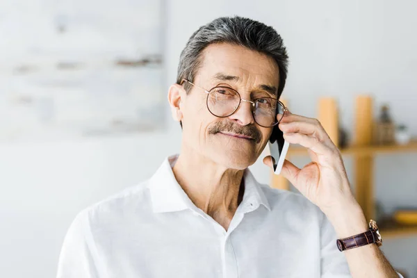 Hombre mayor alegre en gafas hablando en el teléfono inteligente en casa - foto de stock