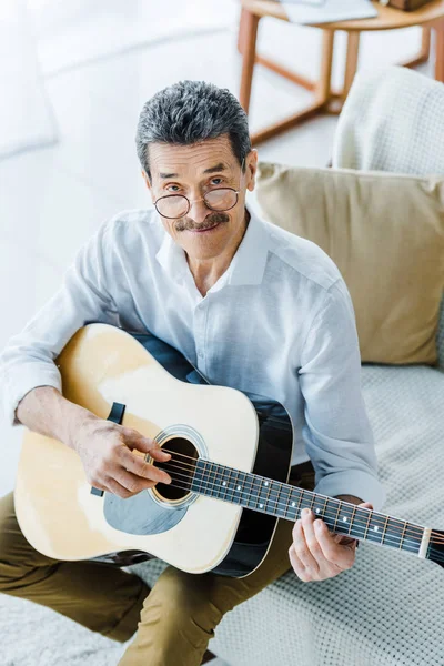 Jubilado alegre en gafas tocando la guitarra acústica en casa - foto de stock