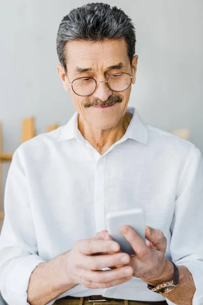 Cheerful senior man in glasses looking at smartphone at home — Stock Photo