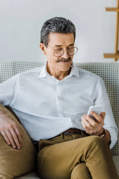 Homem sênior feliz em óculos olhando para o smartphone em casa — Fotografia de Stock