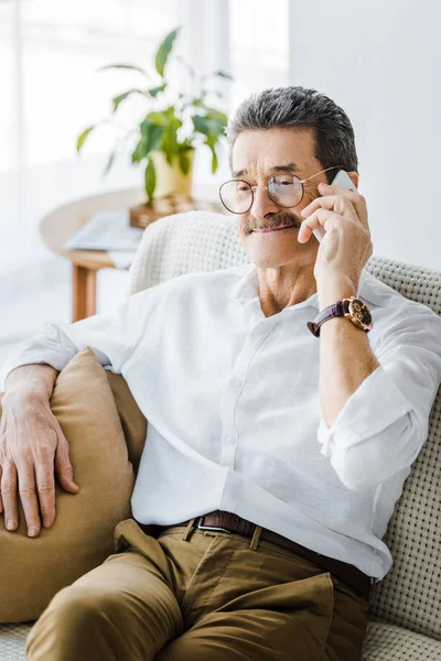 Hombre mayor feliz con bigote hablando en el teléfono inteligente en casa - foto de stock