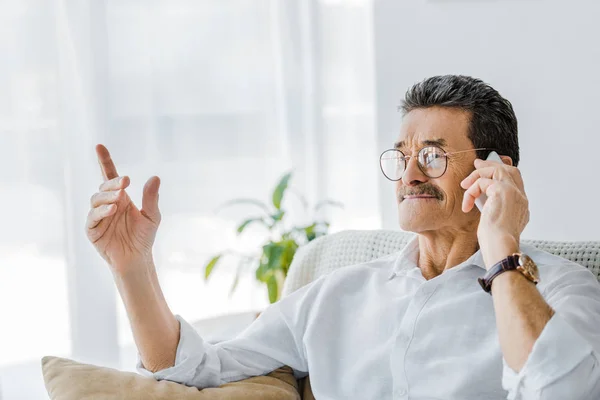 Homem sênior feliz com bigode falando no smartphone e apontando primeiro dedo em casa — Fotografia de Stock