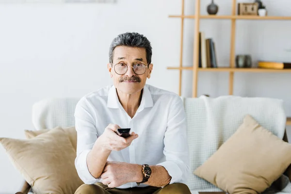 Allegro uomo anziano seduto sul divano e tenendo in mano il telecomando — Stock Photo