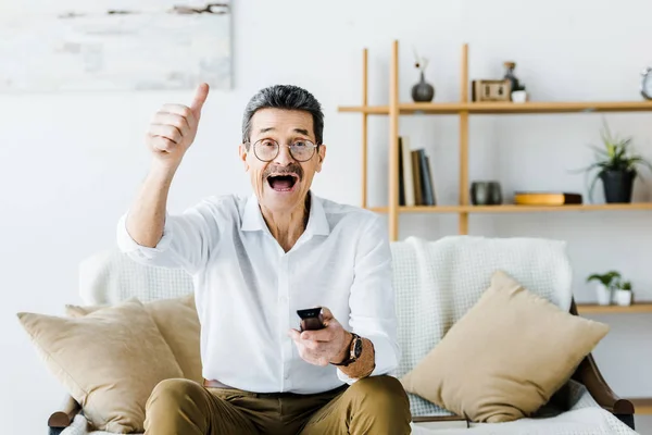 Happy senior man with mustache holding remote control and showing thumb up at home — Stock Photo